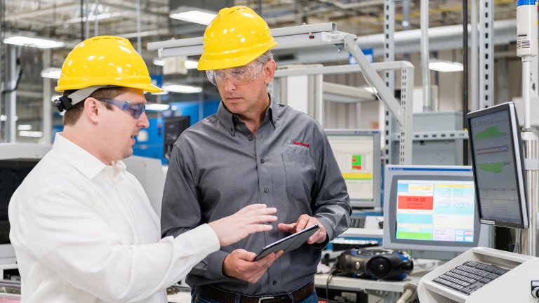 两名工人正在工厂车间环境内通过平板设备协作。一名穿着 Rockwell Automation 衬衫的男性
