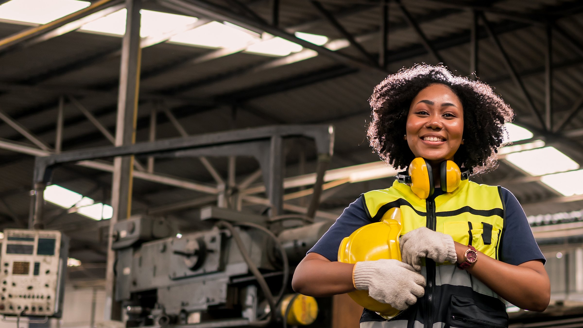 Female engineer in factory setting, dressed with yellow safety vest, headphones and hard hat, standing in front of machinery