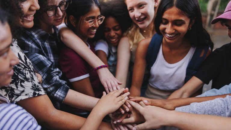 Group of multicultural teenagers smiling cheerfully while putting their hands together in a huddle. Happy young students stacking their hands in unity.